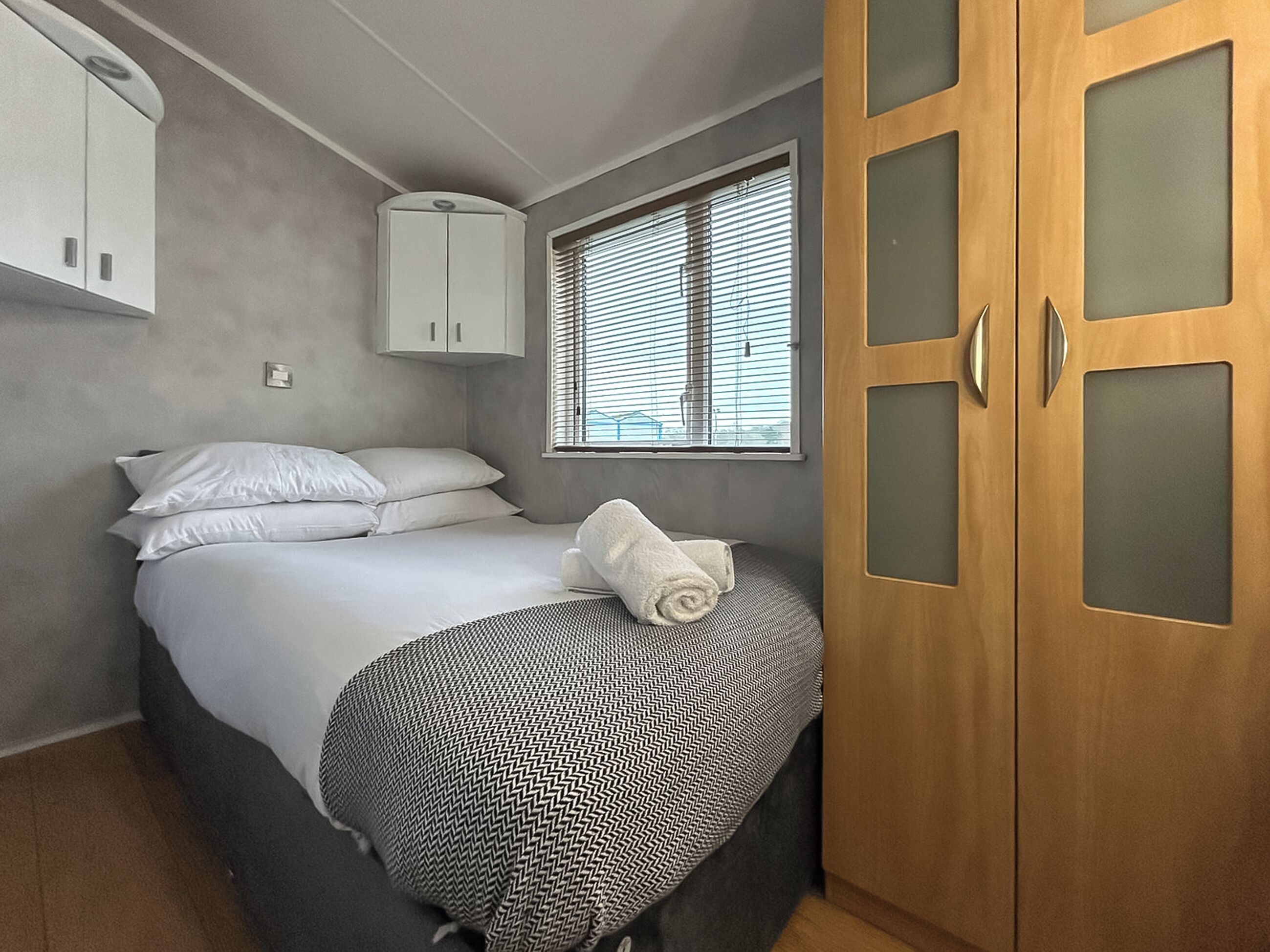 Un dormitorio con cama, ventana con persianas, armario con puertas de vidrio esmerilado y un mueble pequeño.