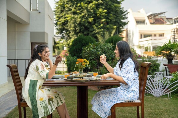 Two people seated at an outdoor table with a vase of flowers, a glass of juice, and plates of food. - Jobner Haus - A Boutique Homestay (Jaipur)