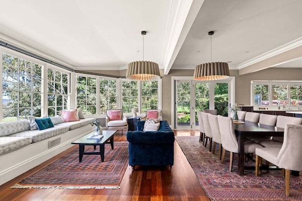 A spacious living room with a large window, a dark blue sofa, a dining area, and a chandelier.