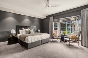 A bedroom with a large bed, a ceiling fan, a window with curtains, and a view of greenery outside.