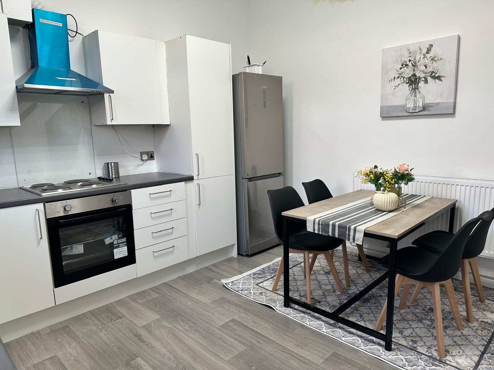 A modern kitchen with white cabinets, a black countertop, a built-in oven, a stainless steel refrigerator, and a dining area with a table and chairs.