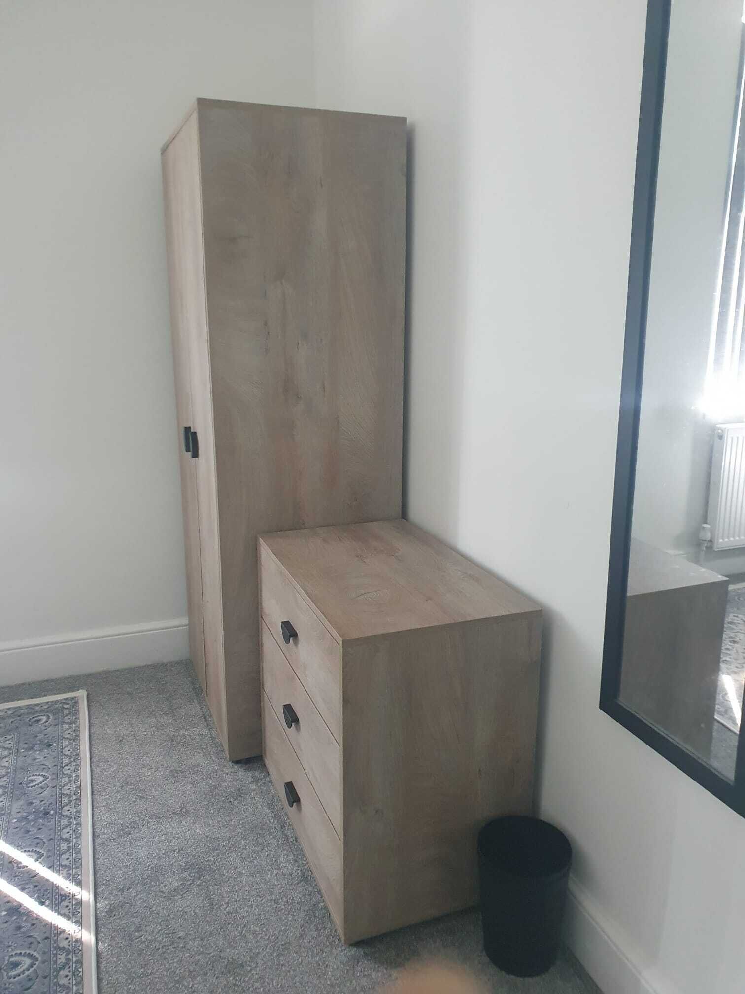 A room with a wardrobe, a small chest of drawers, a black trash bin, and a large mirror on the wall.