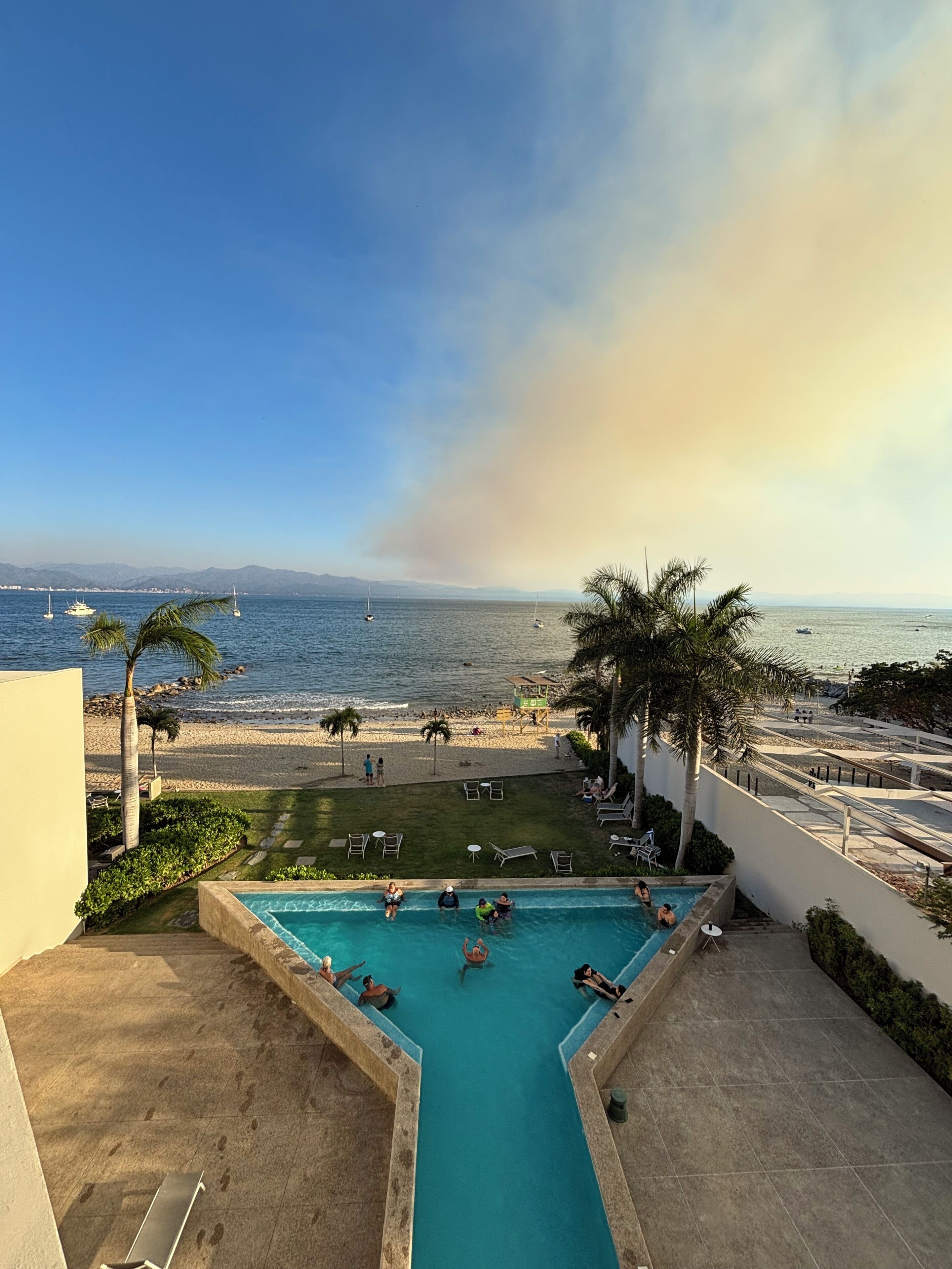 Uma piscina no terraço com vista para a praia e o oceano, rodeada por palmeiras e espreguiçadeiras.