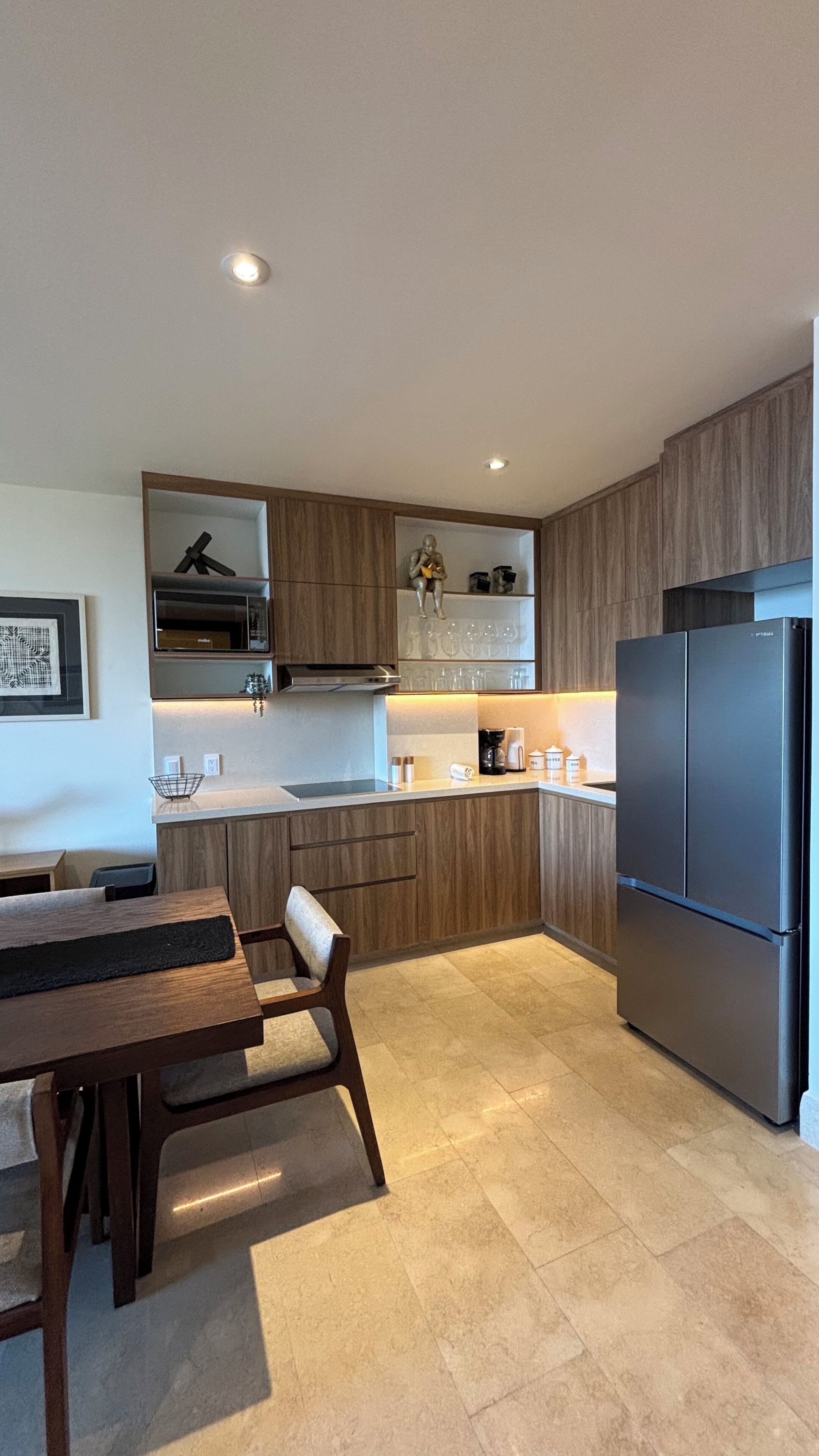 A modern kitchen with wooden cabinets, a stainless steel refrigerator, a dining table with chairs, and a pendant light.