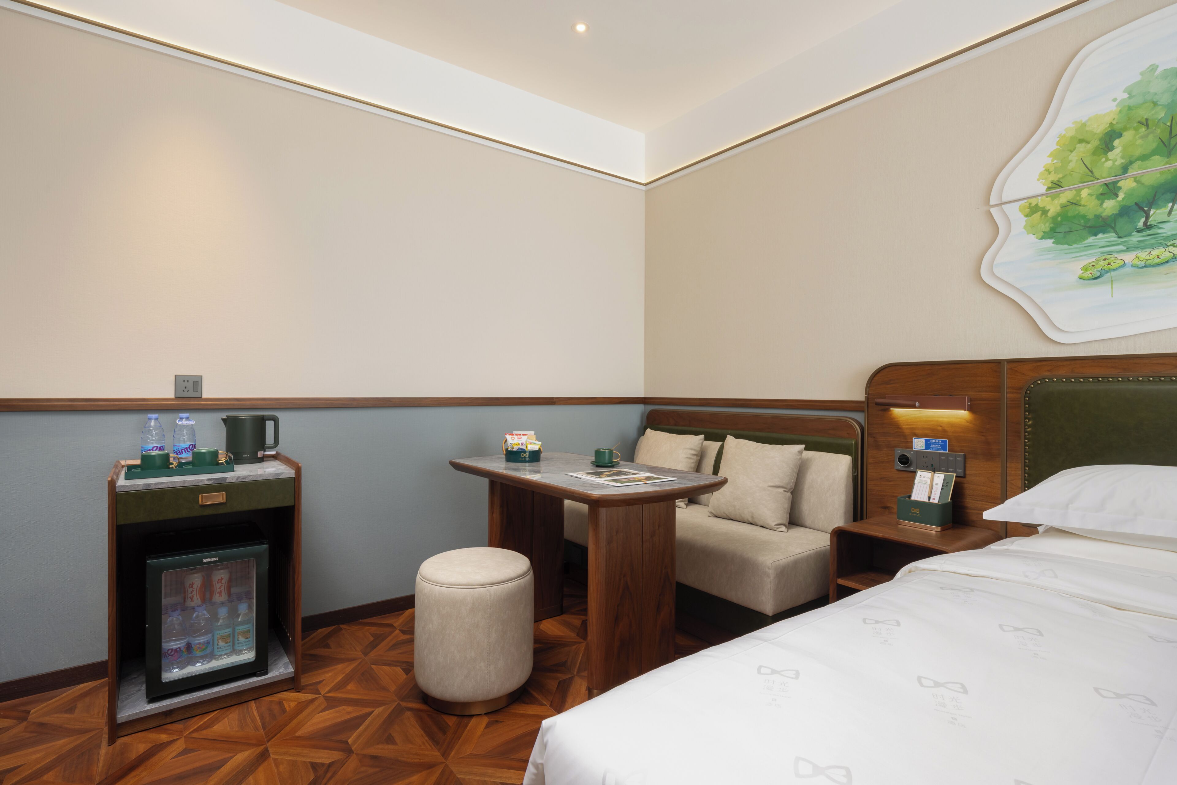 A hotel room with a bed, a small table, a minibar, a seating area, and a painting on the wall.
