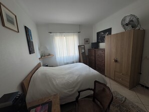 A bedroom with a bed, a wooden wardrobe, a chair, a desk with a computer, a window with curtains, and a wall-mounted fan. - Charming 1-Bedroom Apartment in Alpes-Maritimes (Alpes-Maritimes)