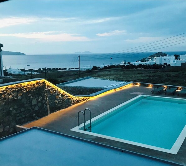 A swimming pool with a stone wall and a view of the sea and white buildings.