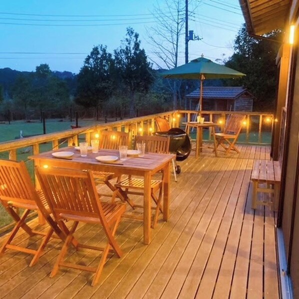 Una terraza de madera con una mesa, sillas, una parrilla y luces colgantes.