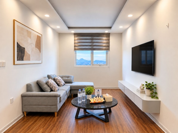 Una sala de estar moderna con un sofá gris, una mesa de centro redonda, un televisor de pantalla plana montado en la pared y una ventana con vistas a montañas.