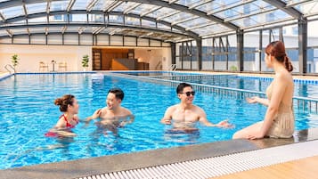 An indoor swimming pool with a glass ceiling, surrounded by lounge chairs and a view of the city skyline.