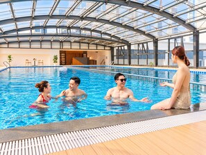 An indoor swimming pool with a glass ceiling, surrounded by lounge chairs and a view of the city skyline.