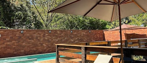 Zona de piscina con muro de ladrillo, terraza de madera y mobiliario exterior.