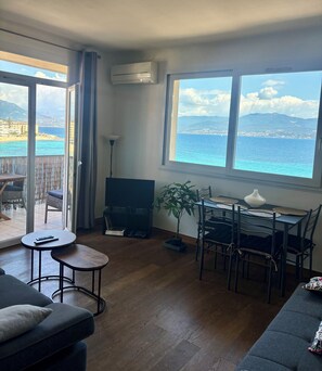 Ein Wohnzimmer mit einer Couch, einem Fernseher, einem Esstisch mit Stühlen, einer Topfpflanze und Blick auf das Meer und die Berge.