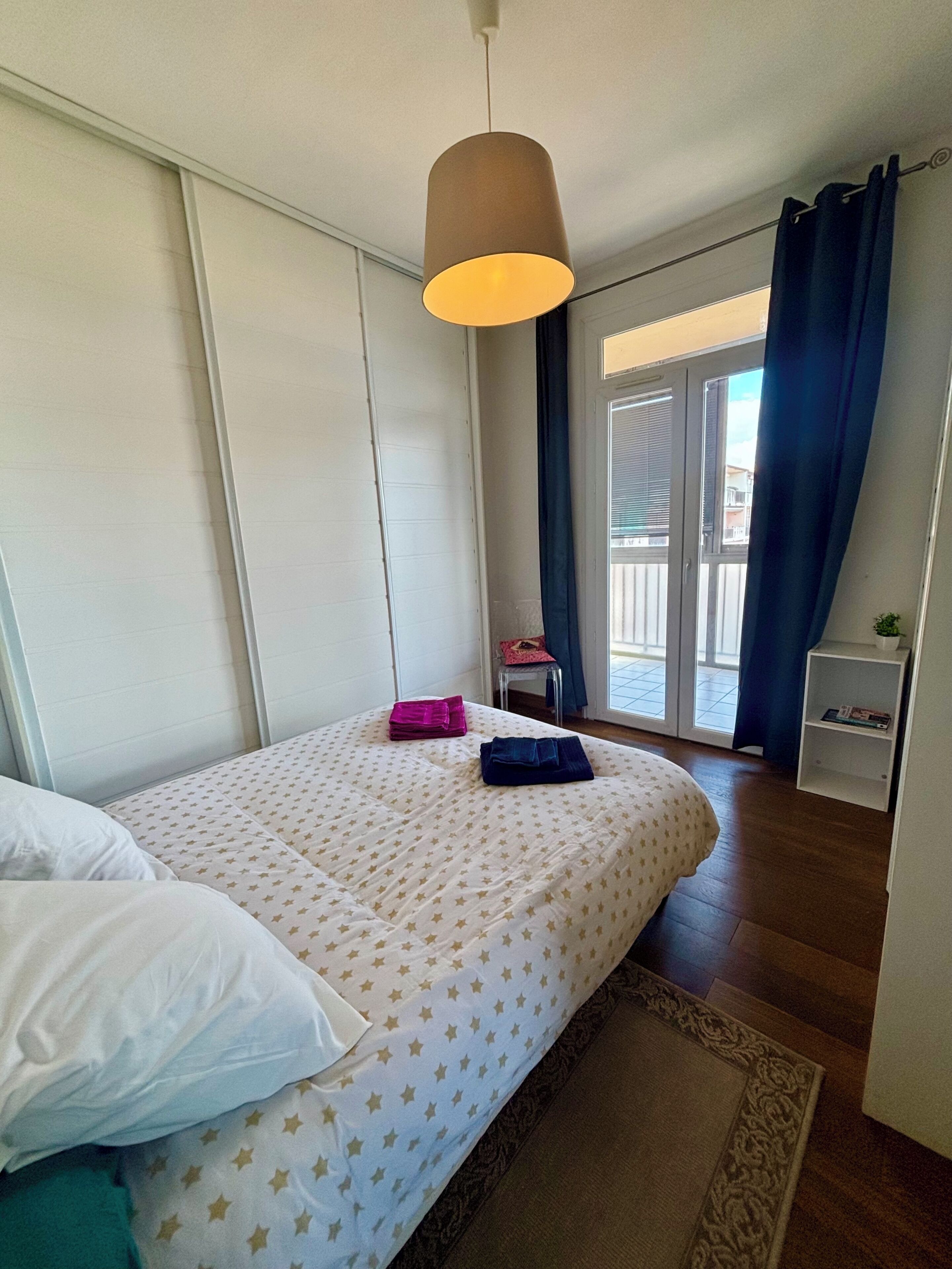 A bedroom with a bed, a hanging light fixture, a window with curtains, a chair, and a small shelf.