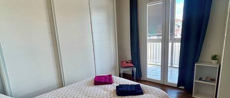 A bedroom with a bed, a hanging light fixture, a window with curtains, a chair, and a small shelf.