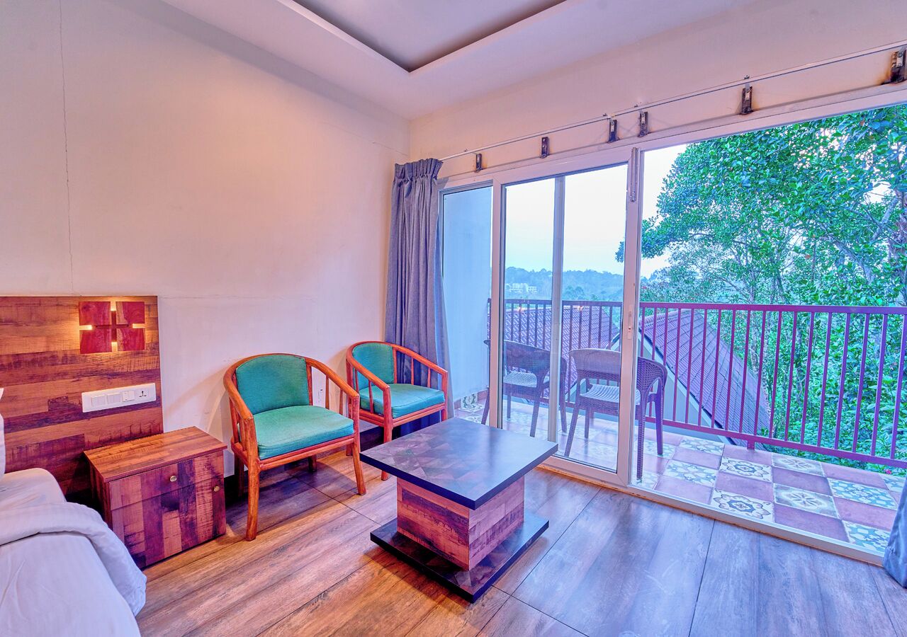 A room with a bed, two chairs, a table, a wooden nightstand, and a balcony with a railing.