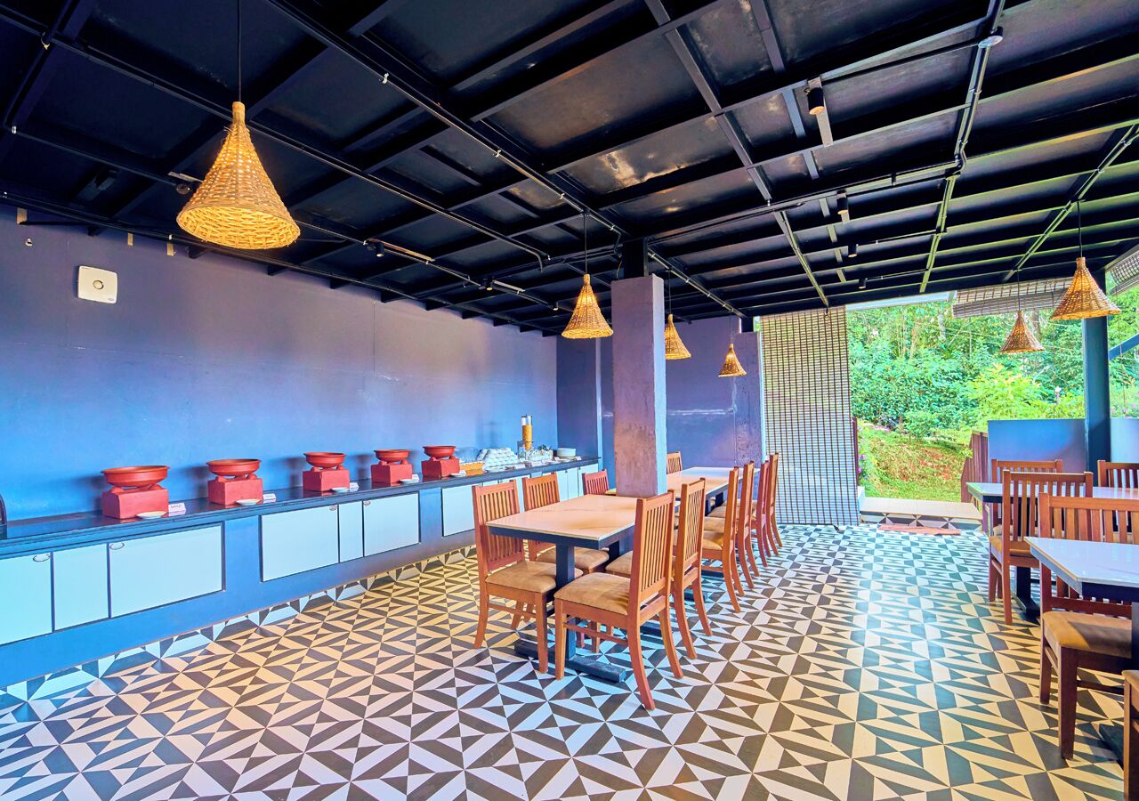 A restaurant with a geometric patterned floor, wooden tables and chairs, hanging wicker lamps, and a view of greenery through a window.