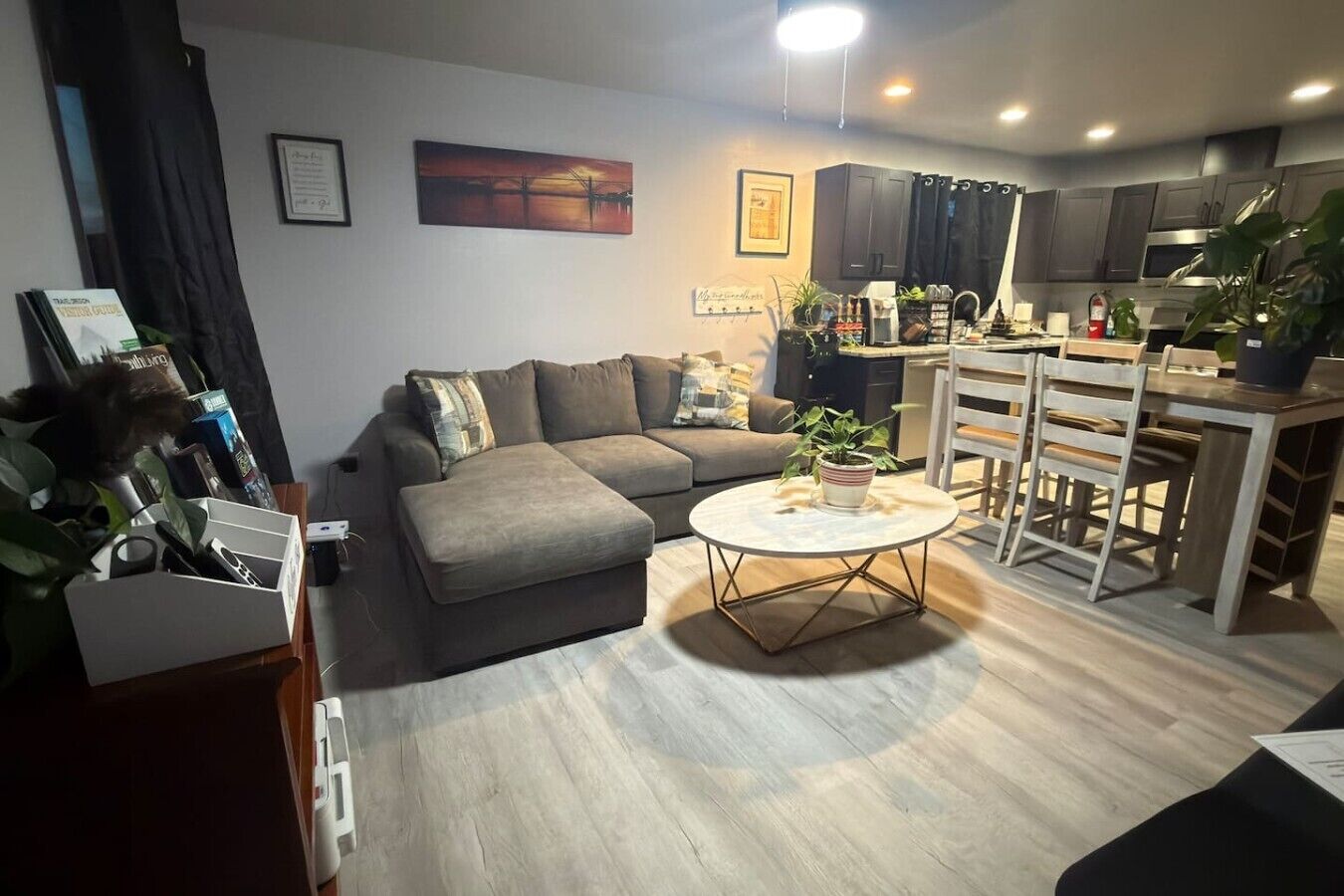 Uma sala de estar moderna com um sofá cinzento, uma mesa de centro redonda e uma área de cozinha com armários escuros e um exaustor de aço inoxidável.