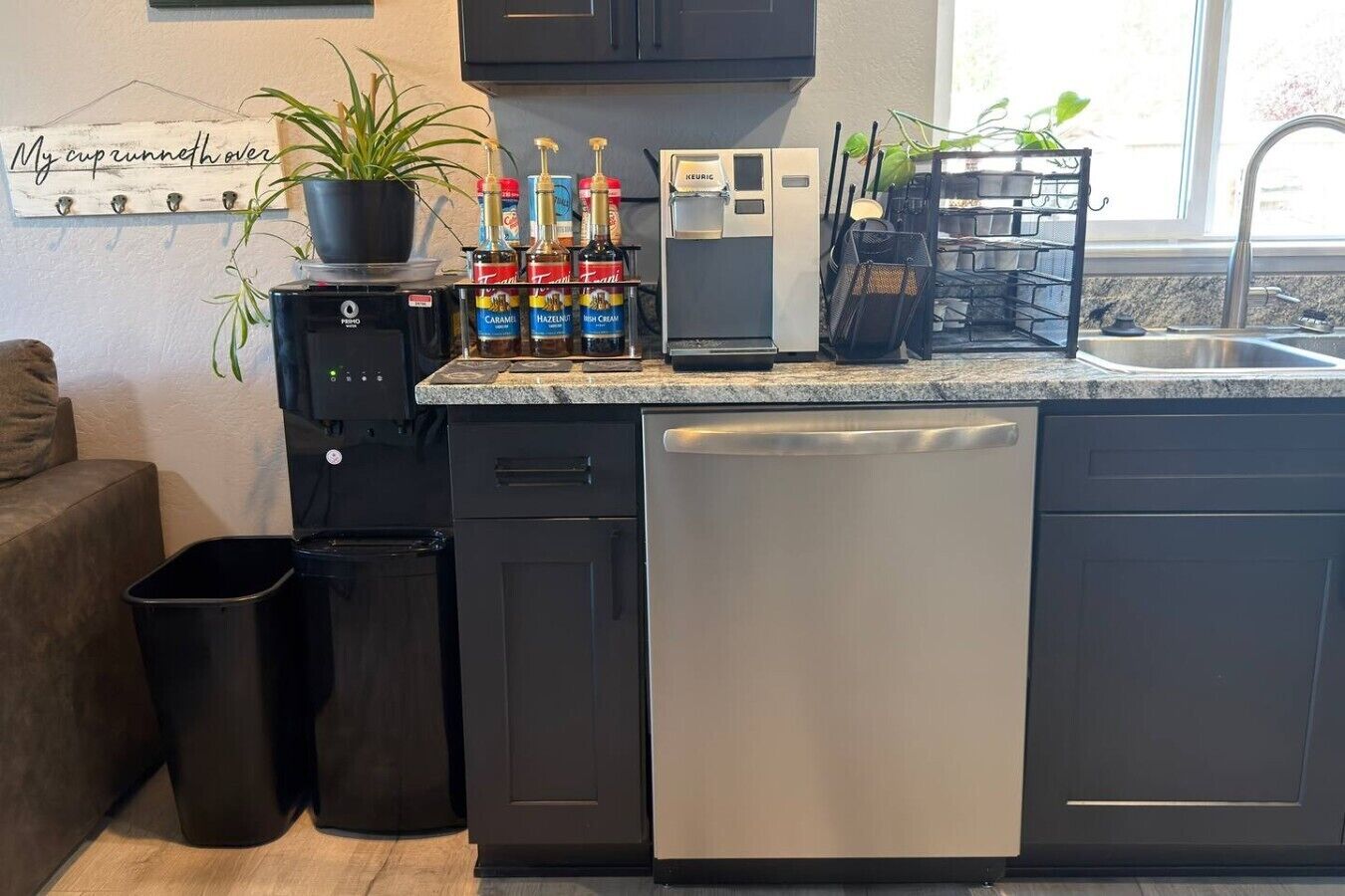Cozinha moderna com máquina de café, chaleira e cafeteira no balcão. Há uma planta em vaso à esquerda e um cesto de lixo sob o balcão.