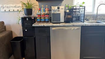 Cozinha moderna com máquina de café, chaleira e cafeteira no balcão. Há uma planta em vaso à esquerda e um cesto de lixo sob o balcão.