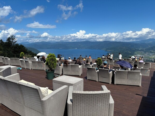 A rooftop terrace with wicker furniture, a potted plant, and a view of a lake and mountains.