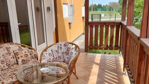 A covered patio with wicker furniture and a glass table, a wooden railing, and a view of a green landscape. - Very nice vacation home with large pool unenclosed property (Balatonszentgyörgy)
