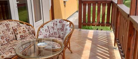 A covered patio with wicker furniture and a glass table, a wooden railing, and a view of a green landscape.