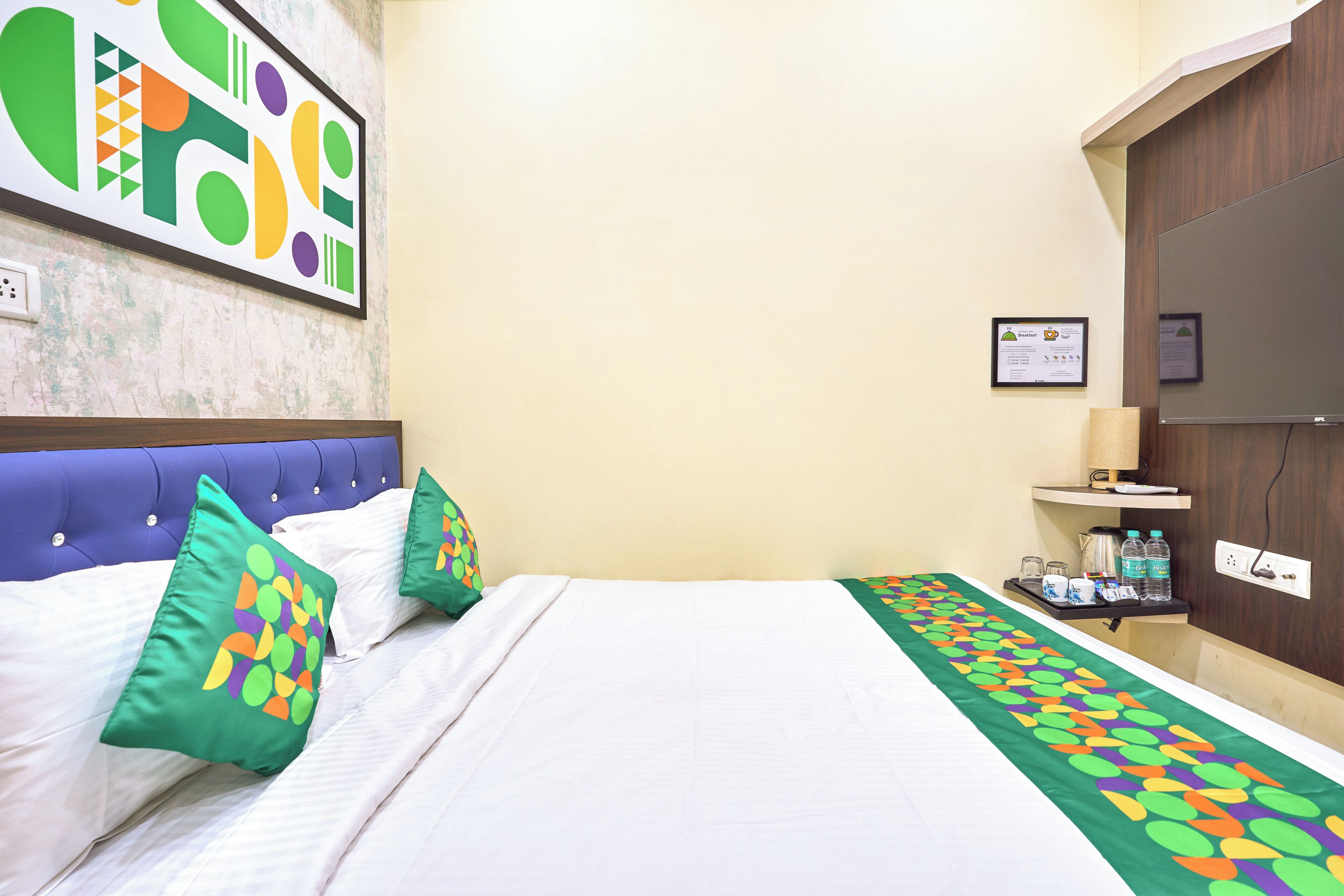Habitació d'hotel amb un llit, almoines, una obra d'art a la paret, una taula de nit amb una làmpada i una ampolla d'aigua.
