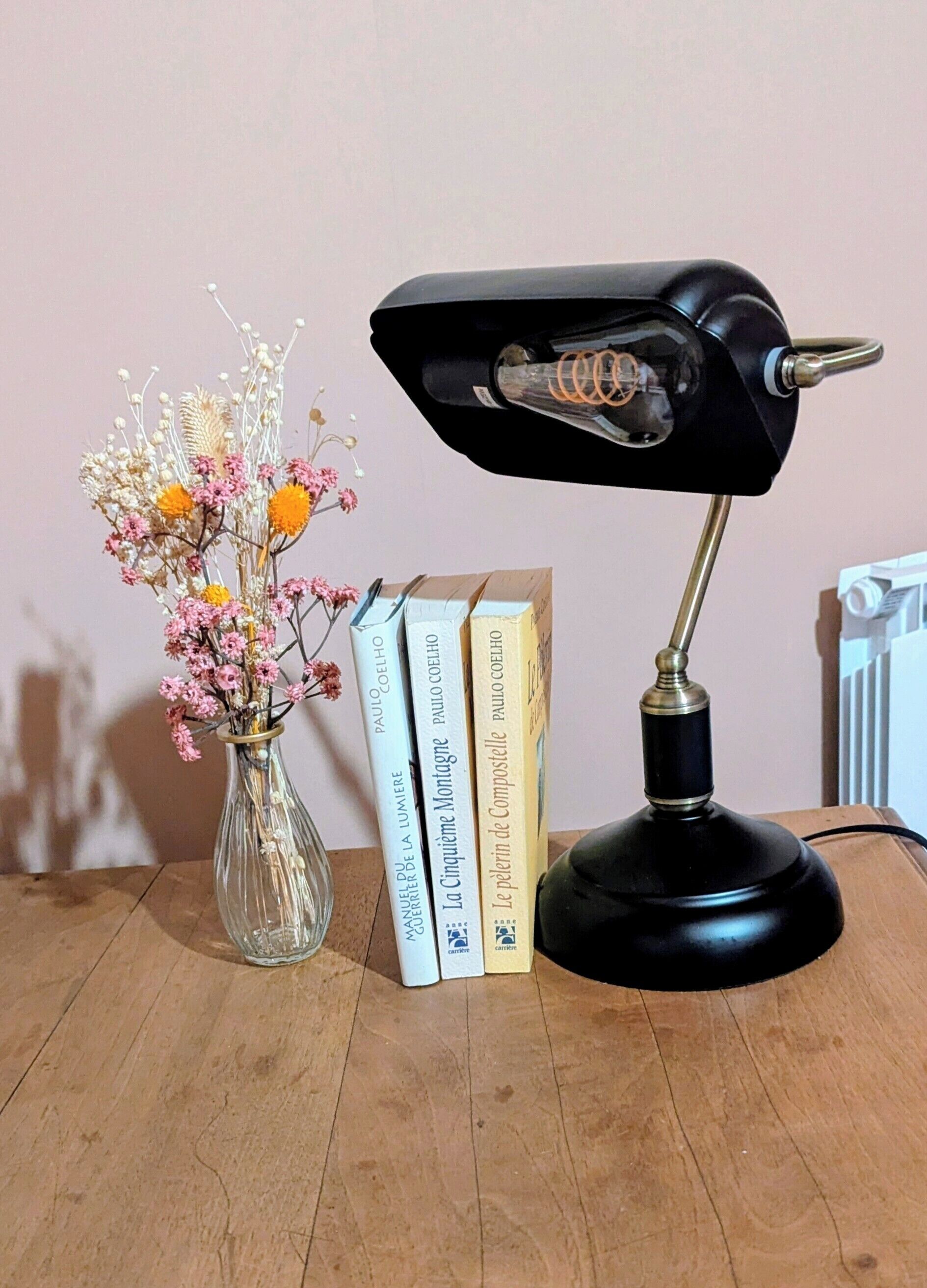 Een bureau met een zwarte bureaulamp, een glazen vaas met gedroogde bloemen en een stapel boeken.