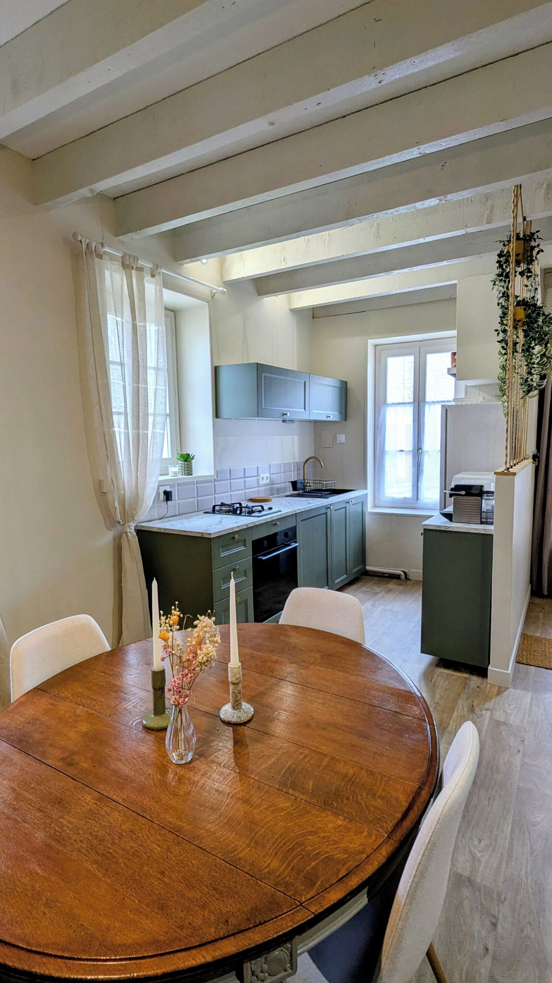 Un coin repas avec une table en bois, des chaises et un vase rempli de fleurs. On aperçoit, au fond, une cuisine avec des placards et une fenêtre.