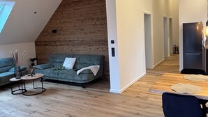 Un espacio habitable moderno estilo loft, con techo inclinado, piso de madera, un sofá gris, una pequeña mesa redonda y una zona de comedor con mesa y sillas de madera.