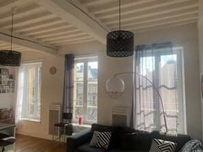 Ein Wohnzimmer mit Sofa, Sessel, Tisch und Bücherregal. Zwei Fenster mit Vorhängen, eine Pendelleuchte und eine Uhr an der Wand.