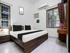 Ein Hotelzimmer mit Bett, Nachttisch, Lampe, Klimaanlage, Fenster mit Vorhängen und einem Kunstwerk an der Wand.