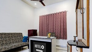 A hotel room with a sofa, a desk with a 'HOTELO' sign, a ceiling fan, and a window with patterned curtains. - Premiumcoastal Inn 3 (Kakinada)