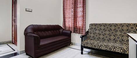 A room with a dark brown sofa, a patterned armchair, a red patterned curtain, and a light-colored tiled floor.
