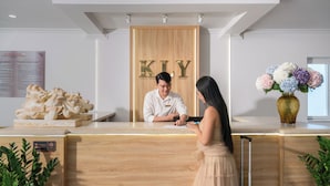 A reception desk with a wooden panel displaying 'KLY' in gold letters, a receptionist, a guest, and a vase with flowers. - KLY Hotel & Retreat (Ho Chi Minh City)