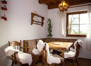 Un coin repas avec une table et des chaises en bois, un lustre, une fenêtre avec des rideaux et une étagère murale ornée de pots.