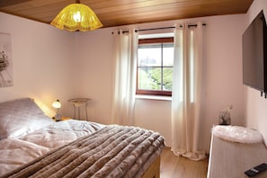 A bedroom with a bed, a bedside lamp, a window with curtains, a small table with a vase, and a mounted TV. - Cottage 'Lieblingsplatz Am Bach' with Wi-Fi (Windorf)