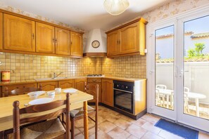 A kitchen with wooden cabinets, a dining table with chairs, a clock, and a view through a glass door. - La Brise de Valras – Air-Conditioned House (Valras-Plage)
