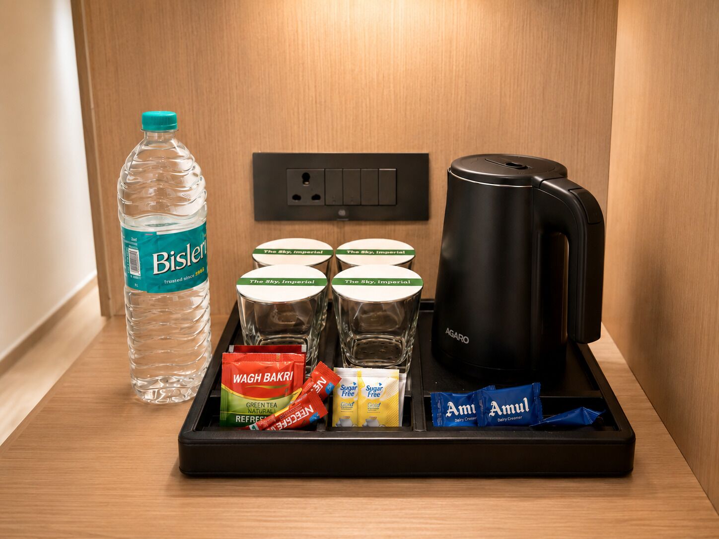 A minibar setup with a bottle of Bisleri water, four glasses labeled 'The Sky, Imperial,' a black coffee maker, and various food items including green tea, sugar-free packets, and dairy creamers.