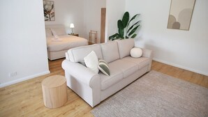 Ein modernes Wohnzimmer mit einer Couch, einem runden Holzschemel, einer großen Pflanze und einem Bett mit einem Nachttisch.