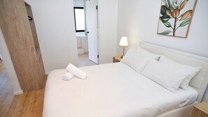 Ein ordentlich bezogenes Bett mit weißen Leinen, ein Nachttisch aus Holz mit einer Lampe und ein an der Wand befestigtes Kunstwerk.