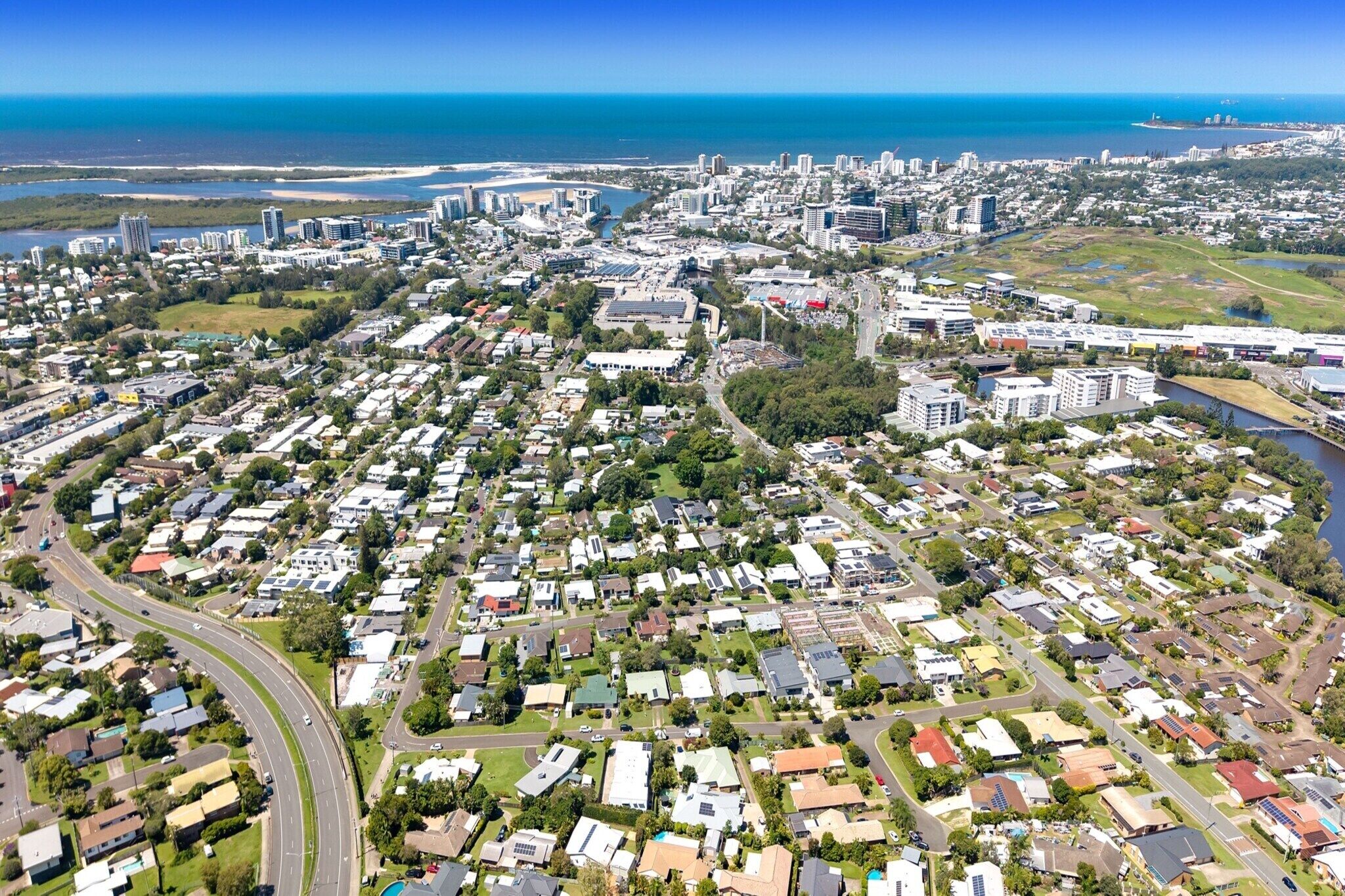 An aerial view of a coastal city with dense residential areas, a prominent highway, and a large body of water in the background.