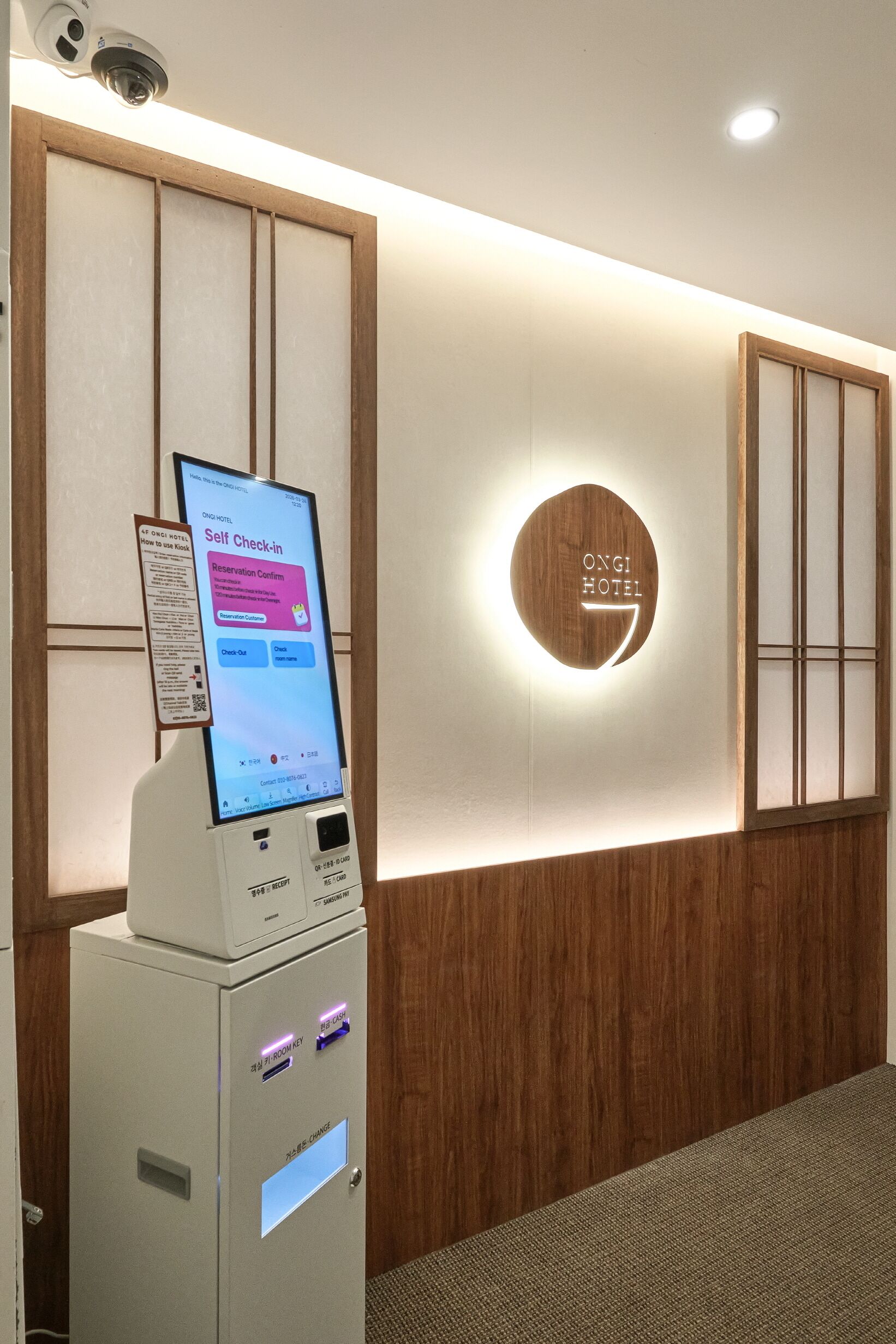 A self-check-in kiosk, a wooden wall panel, and a circular sign with 'ONGI HOTEL' on a modern interior.