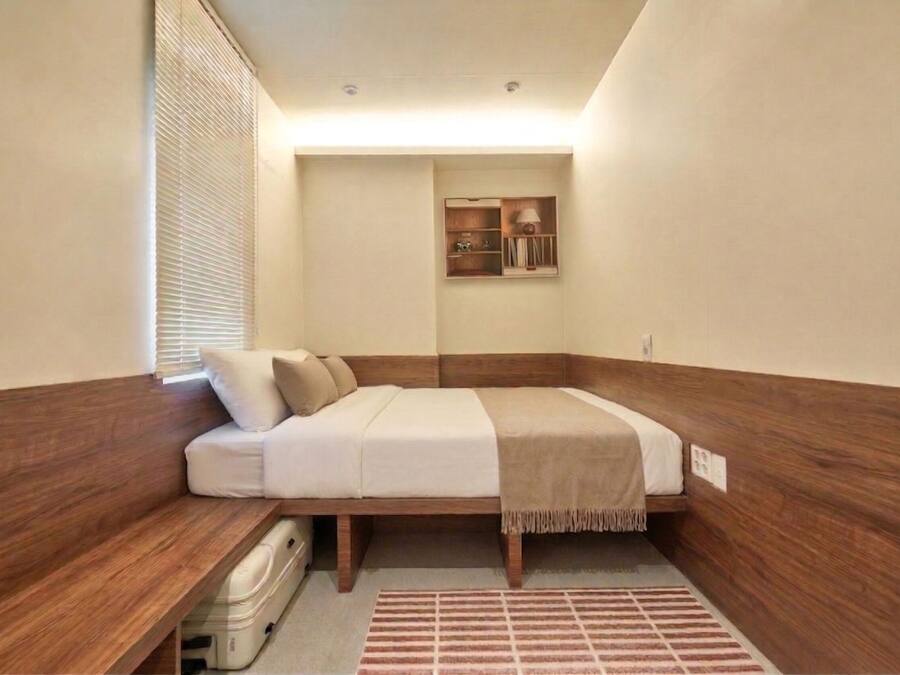 Une petite chambre à coucher, bien rangée, avec un lit, une table de chevet et une étagère fixée au mur, sur laquelle se trouve une lampe.