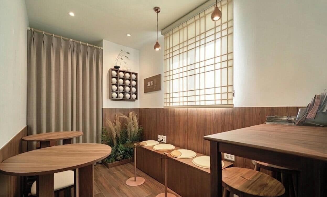 A restaurant interior with wooden tables and chairs, a window with a grid pattern, and a wall-mounted shelf with round objects.