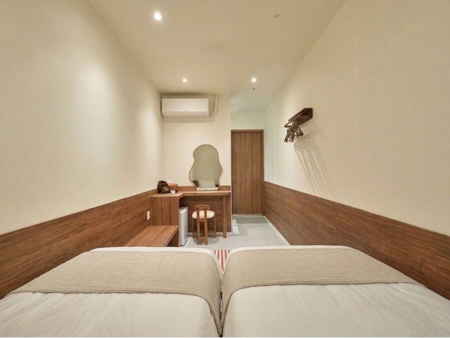 Habitación de hotel con dos camas, un escritorio, una silla, una mesita y un estante en la pared.