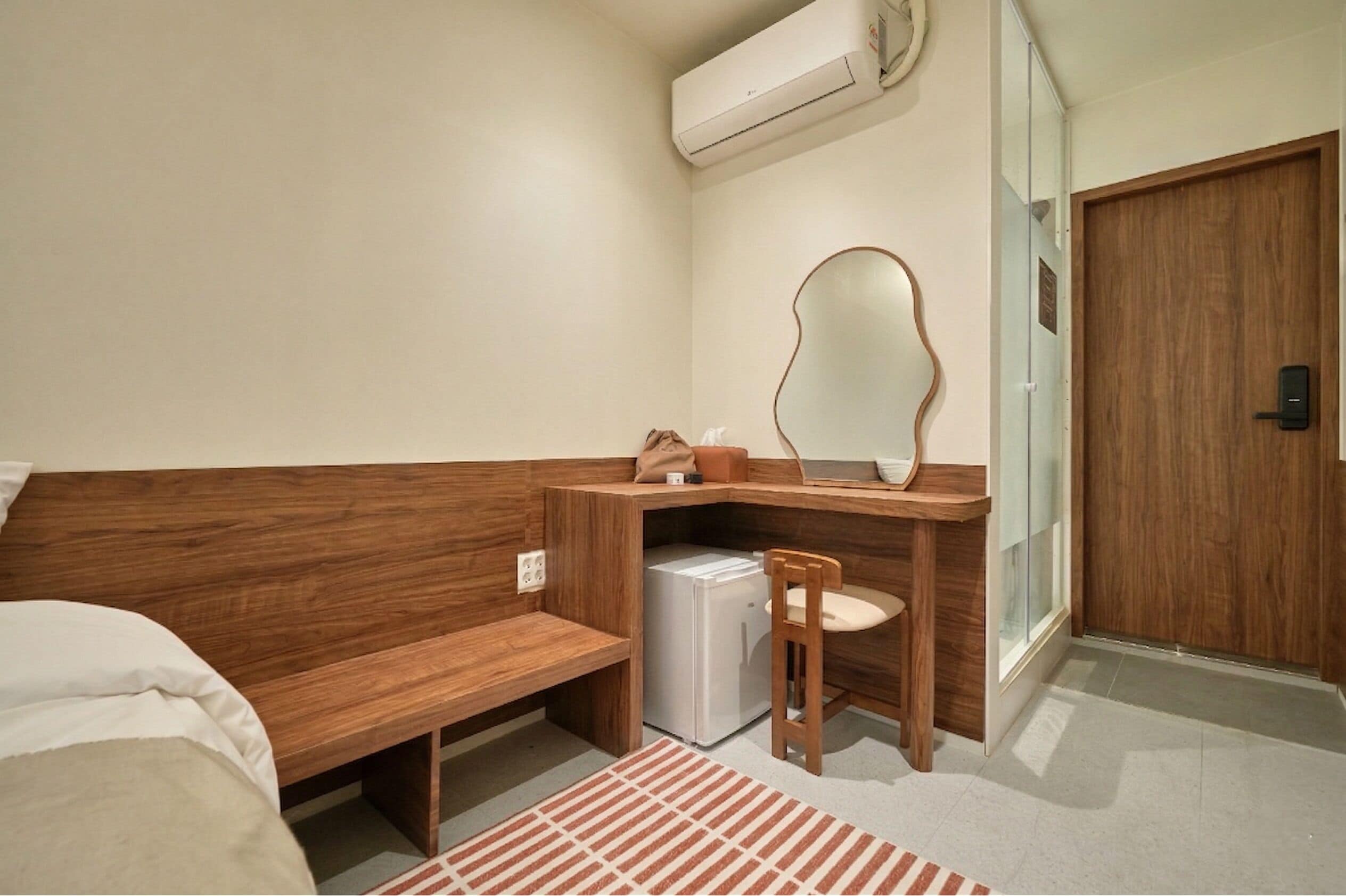 Quarto de hotel compacto com um banco de madeira, uma pequena escrivaninha com espelho, um frigorífico branco e uma porta de correr de vidro.