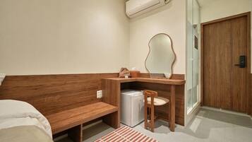 Quarto de hotel compacto com um banco de madeira, uma pequena escrivaninha com espelho, um frigorífico branco e uma porta de correr de vidro.