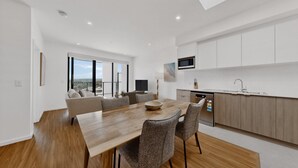 A modern kitchen with white cabinets, a wooden dining table, and a built-in oven. - Brand New 2 Bed Apartment with City Views (Cannington)